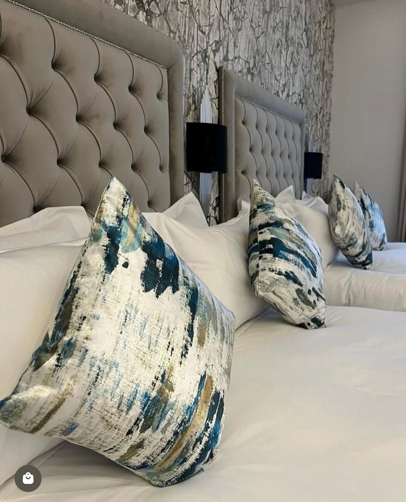 May include: A bedroom scene featuring two upholstered headboards with button tufting. White bedding is topped with decorative pillows in shades of blue, teal, and gold. Black lamps are mounted on the wall between the headboards. The wallpaper has a gray and white pattern.