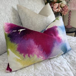 May include: A rectangular throw pillow featuring a vibrant watercolor-style print with blended splashes of magenta, deep purple, lime green, and sky blue. Behind it sits a second, smaller throw pillow with a neutral light gray herringbone pattern.