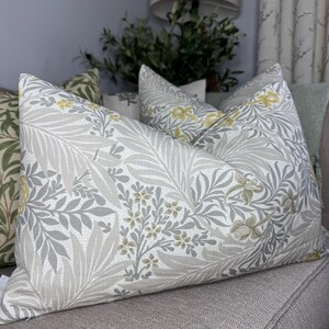 Könnte beinhalten: Dekorative Kissen mit einem botanischen Muster. Die Kissen haben einen hellgrauen Hintergrund mit einem sich wiederholenden Design aus grauen Blättern und gelben Blüten. Die Kissen liegen auf einem grauen Sofa.