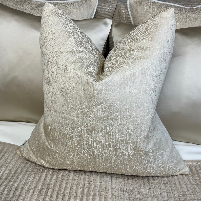 May include: A soft, textured, beige throw pillow with a subtle shimmer. The pillow is made of a luxurious fabric and has a square shape.