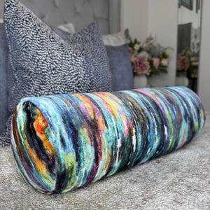 May include: A cylindrical bolster pillow with a vibrant abstract design. The pillow features a blend of colors including blue, green, orange, and purple, creating a watercolor effect. The pillow is placed on a bed with other pillows in the background.