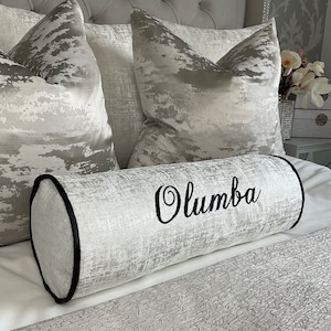 May include: A white and silver cylindrical pillow with a black embroidered name "Olumba" on a bed with matching pillows.