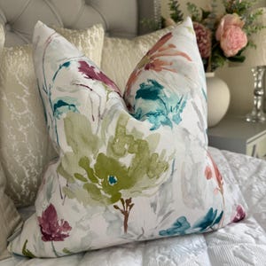 May include: A decorative throw pillow with a floral pattern. The pillow features a white background with watercolor-style flowers in shades of green, teal, pink, and purple. The pillow is placed on a white quilted bedspread.