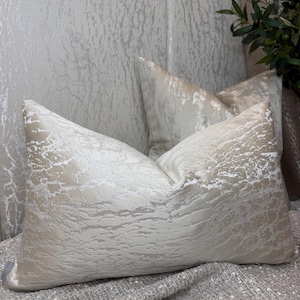 May include: Two rectangular throw pillows featuring a light beige and cream color palette with a textured, marbled, or cracked surface pattern. The fabric has a subtle sheen, and the pillows are arranged with one positioned in front of the other.