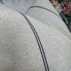 May include: Close-up of a folded, beige linen fabric with two dark blue stripes running parallel. The fabric has a textured weave, and the stripes add a contrasting design element. The overall aesthetic is rustic and natural.