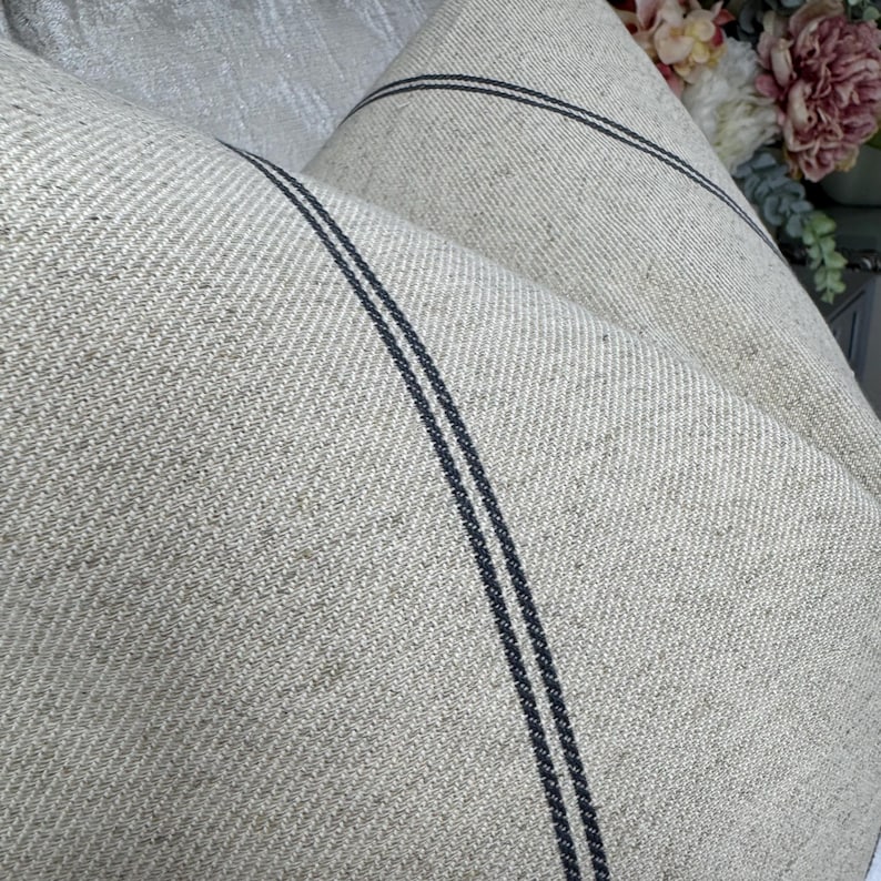 May include: Close-up of a folded, beige linen fabric with two dark blue stripes running parallel. The fabric has a textured weave, and the stripes add a contrasting design element. The overall aesthetic is rustic and natural.