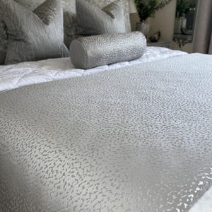 Peut inclure: Un lit avec une palette de couleurs grises et blanches. Le lit a une tête de lit grise et est recouvert d'un couvre-lit matelassé blanc. Des oreillers décoratifs gris et un traversin sont disposés sur le lit. Le chemin de lit est en tissu gris texturé.