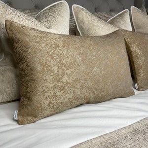 Pode incluir: Um conjunto de quatro almofadas decorativas com um tecido texturizado dourado metálico. As almofadas estão dispostas em uma cama branca com uma colcha estampada.