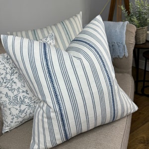 May include: Two decorative throw pillows with a white background and blue stripes. One pillow has a floral pattern in blue and white. The pillows are on a light brown couch.