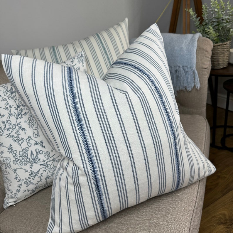 May include: Two decorative throw pillows with a white background and blue stripes. One pillow has a floral pattern in blue and white. The pillows are on a light brown couch.