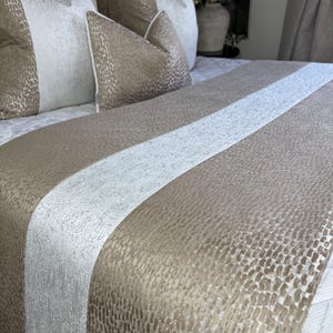 Puede incluir: Un camino de cama y almohadas en un dormitorio. El camino de cama y las almohadas presentan un patrón texturizado y moteado en tonos beige y crema. Una franja central de color crema recorre la longitud del camino de cama. Las almohadas tienen un diseño similar.
