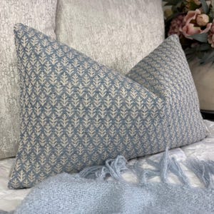 Op de afbeelding: Een rechthoekig sierkussen met een herhalend blauw en crèmekleurig geometrisch patroon van kleine, gestileerde boomvormen. Het kussen heeft een textielafwerking met textuur en is geplaatst voor een lichtgekleurd kussen met een subtiel patroon.