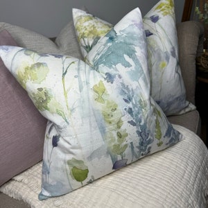 May include: Three decorative throw pillows with a watercolor floral pattern in shades of green, blue, and purple. The pillows are arranged on a white couch.