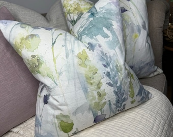 May include: Three decorative throw pillows featuring a watercolour floral design in shades of green, blue and purple. The pillows are arranged on a white sofa.