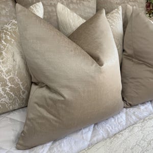 May include: A selection of beige and cream-coloured decorative cushions. The cushions have textured and smooth surfaces, with a focus on the soft, neutral tones. The arrangement suggests a cosy and inviting home decor setting.