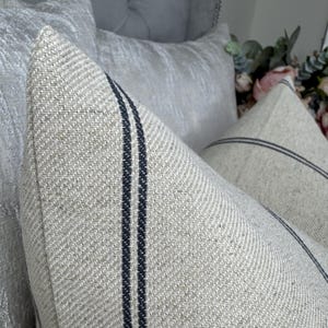 May include: Close-up of two decorative throw pillows. The pillows are a light beige color with two dark blue vertical stripes. The pillows have a textured fabric and are placed in front of other light-colored pillows.