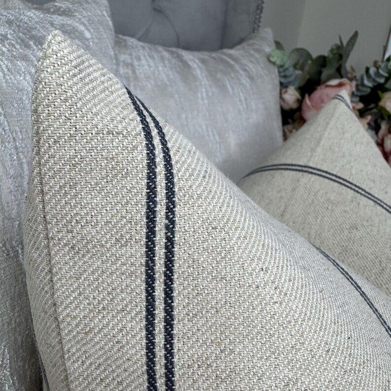 May include: Close-up of two decorative throw pillows. The pillows are a light beige color with two dark blue vertical stripes. The pillows have a textured fabric and are placed in front of other light-colored pillows.