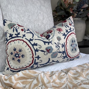 May include: Embroidered rectangular cushion with a cream background and floral patterns in navy blue, burgundy, and light blue. The cushion is placed on a white bed, with a beige patterned throw visible at the bottom.