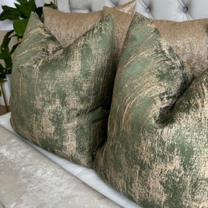 May include: Two green and gold throw pillows with a distressed, textured fabric. The pillows are on a white bed with a light grey blanket.