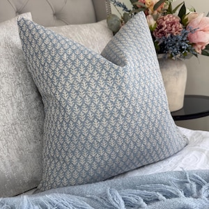 May include: A square throw pillow featuring a light blue fabric base with a repeating white botanical pattern of small, stylized leaf sprigs. The pillow has a textured appearance and is positioned upright, showcasing a clean, structured edge design.