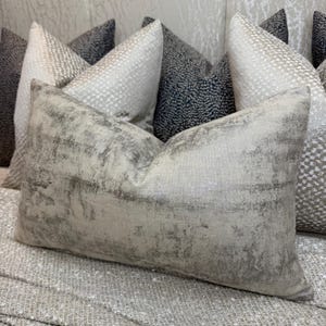 Puede incluir: Un cojín rectangular de tela beige claro con un acabado metálico plateado desgastado. El cojín tiene una apariencia texturizada con vetas horizontales de color gris oscuro y plata, creando un efecto desgastado y moteado en toda la superficie.