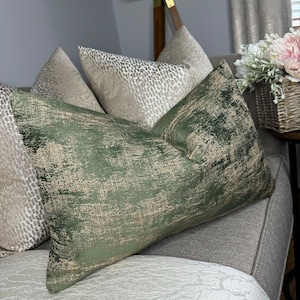 May include: Three decorative throw pillows, one green with gold accents, one beige with a subtle pattern, and one beige with a more pronounced pattern. The pillows are arranged on a grey sofa.