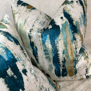 May include: Three decorative throw pillows with a white background and a bold abstract design in shades of teal, blue, and gold. The pillows have a soft, textured appearance, suggesting a comfortable and stylish home decor item.