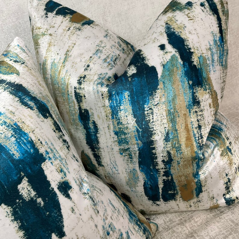 May include: Three decorative throw pillows with a white background and a bold abstract design in shades of teal, blue, and gold. The pillows have a soft, textured appearance, suggesting a comfortable and stylish home decor item.