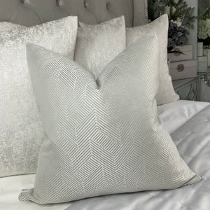 May include: Three decorative throw pillows with a white and silver geometric pattern. The pillows are arranged on a bed with a white and silver bedspread.