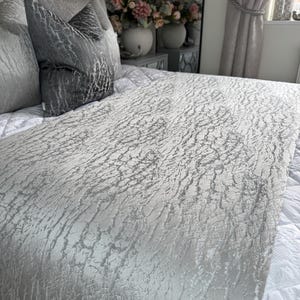 Puede incluir: Una cama con cabecero acolchado, con una colcha plateada texturizada y almohadas a juego. La habitación está decorada en tonos neutros, con arreglos florales y cortinas visibles en el fondo.