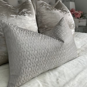 Puede incluir: Una colección de cojines decorativos en tonos grises y plateados. El cojín delantero tiene un patrón texturizado y repetitivo. Los otros cojines tienen un diseño similar a una nube. Los cojines están colocados sobre una colcha blanca.