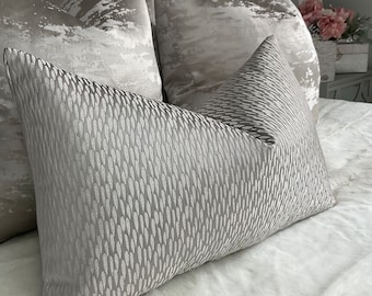 May include: A collection of decorative pillows in shades of gray and silver. The front pillow has a textured, repeating pattern. The other pillows have a cloud-like design. The pillows are arranged on a white bedspread.