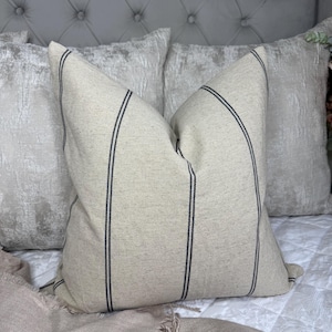 May include: A beige throw pillow with two vertical navy blue stripes. The pillow has a textured fabric and is placed on a bed with other pillows. The pillow is a decorative item for a bedroom.