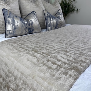 May include: A bed is made with a beige textured bedspread and several decorative pillows. The pillows have a silver and blue abstract design with navy blue trim. The bed is made with a white quilted bed skirt.