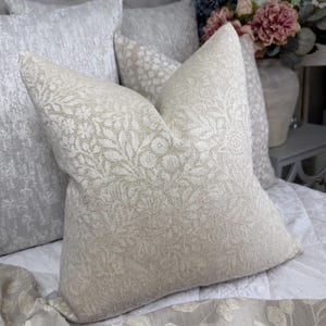 May include: Several decorative cushions in cream and beige tones. The front cushion has a detailed floral design. Other cushions have textured or patterned surfaces. The cushions are arranged on a white bedspread.