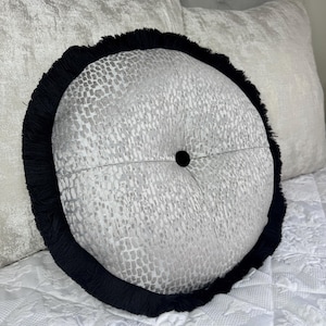 以下が含まれることがあります： 黒のフリンジの縁取りが付いた、グレーと白の模様の丸いクッション。クッションの中央にボタンが付いている。