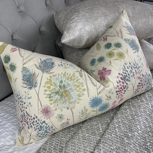 Puede incluir: Dos almohadas decorativas con patrones florales. Una almohada tiene un fondo crema con flores azules, verdes y rosas. La otra almohada tiene un fondo plateado con un brillo sutil.