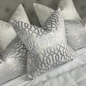 May include: A set of four decorative throw pillows with a geometric pattern in shades of silver and white. The pillows are arranged on a white bedspread.