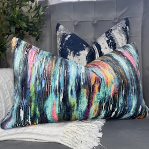 May include: Decorative lumbar pillow with a vibrant abstract design in shades of blue, green, pink, and yellow. A second pillow with a navy and white pattern is visible in the background. The pillows are on a gray upholstered surface.
