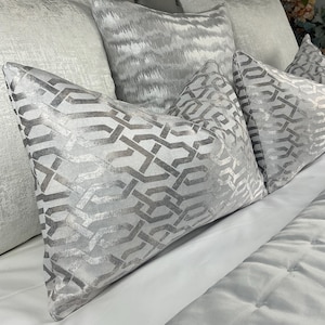 May include: A collection of decorative throw pillows in shades of gray and silver. The pillows feature various textures and patterns, including a geometric design and a wavy pattern. The pillows are arranged on a white bedspread, creating a luxurious and elegant aesthetic.