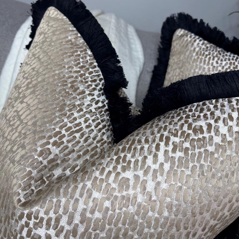 K&ouml;nnte beinhalten: Zwei dekorative Kissen mit einem Schlangenhautmuster in Beige und Gold. Die Kissen haben einen schwarzen Fransenbesatz an den R&auml;ndern.