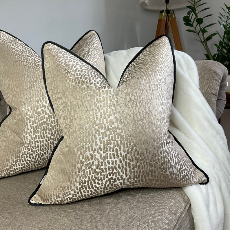 May include: Two decorative throw pillows with a beige and brown abstract pattern. The pillows have a black trim around the edges.