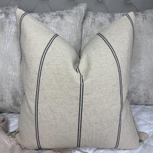 May include: A beige throw pillow with two vertical black stripes. The pillow is on a white quilted bedspread, with a light pink blanket and two other pillows in the background. The pillow has a textured appearance.