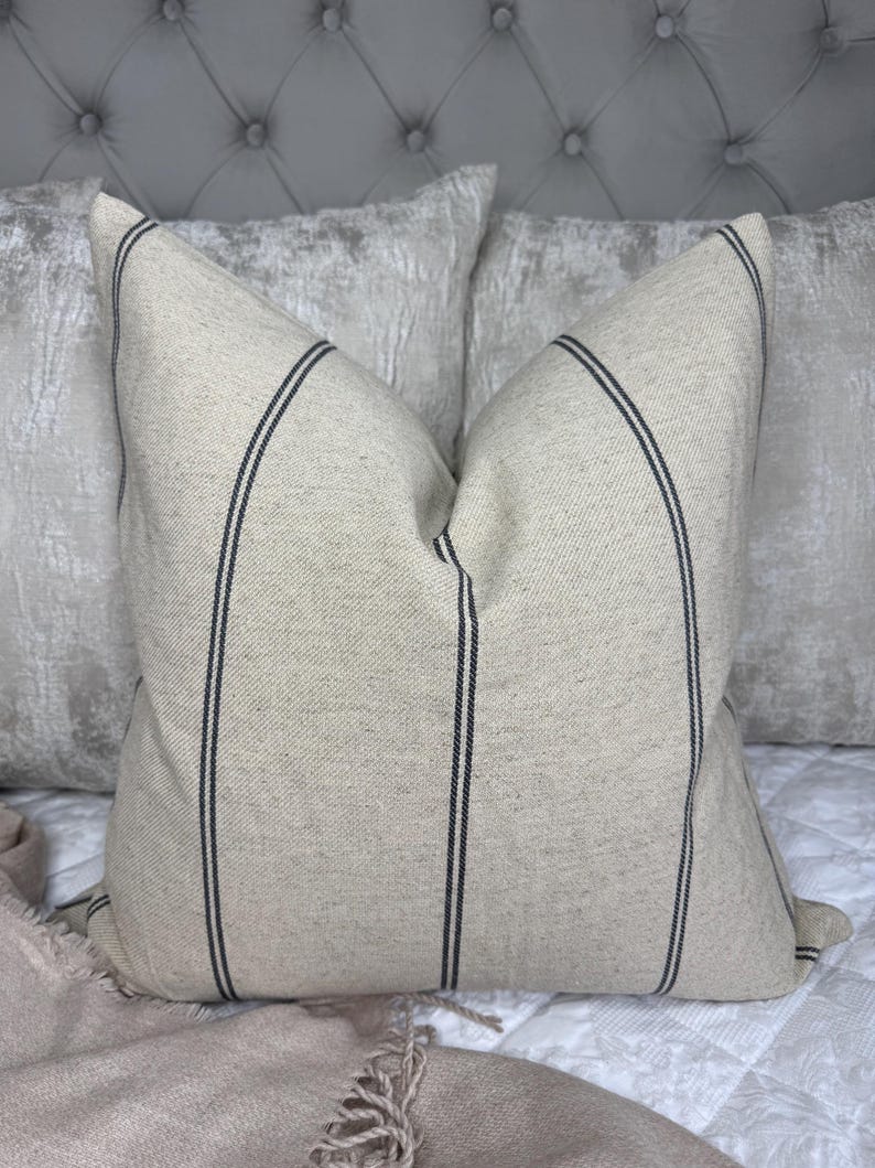 May include: A beige throw pillow with two vertical black stripes. The pillow is on a white quilted bedspread, with a light pink blanket and two other pillows in the background. The pillow has a textured appearance.