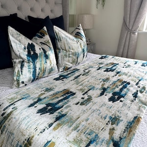 May include: A bed with a gray tufted headboard, navy blue pillows, and decorative pillows with a white, teal, and gold abstract design. A matching bed runner with the same pattern is visible. The bed is made up with white bedding.