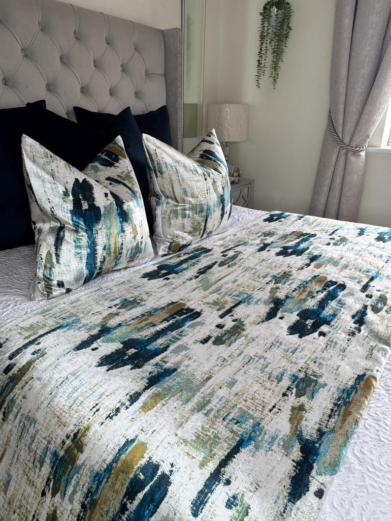 May include: A bed with a gray tufted headboard, navy blue pillows, and decorative pillows with a white, teal, and gold abstract design. A matching bed runner with the same pattern is visible. The bed is made up with white bedding.