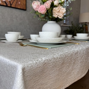 May include: A white tablecloth with a textured, crackled finish. The tablecloth is set with white plates, bowls, and cups. A bouquet of pink flowers in a white vase is on the table.