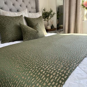 Op de afbeelding: Een groene bedsprei met een gouden stippenpatroon. De sprei ligt op een bed met groene en witte kussens.