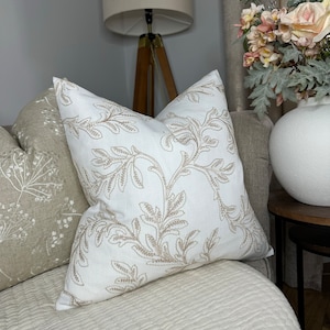 May include: A white throw pillow with a beige floral embroidery design. The pillow is on a white couch with a beige throw pillow behind it.