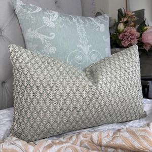 May include: Two decorative pillows on a bed. The front pillow has a green and white leaf pattern. The back pillow is light blue with a white floral design. The pillows are on a white bedspread with a patterned throw.